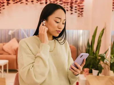 A young woman wearing wireless earbuds looks at her smartphone.