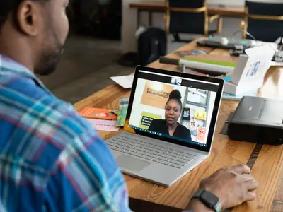 A man in a plaid shirt talks on a video call at his laptop.
