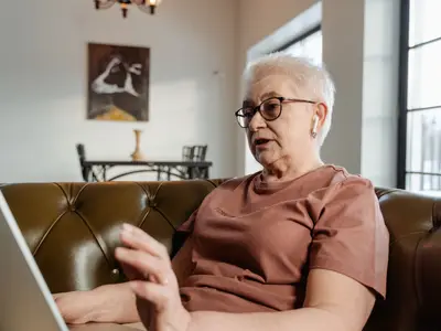 An older woman sits on a couch using a laptop.