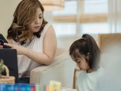 A mother holds her phone while looking at her young daughter at home.