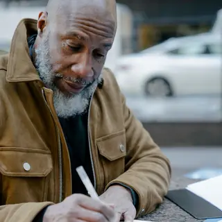 An older Black man with a gray beard writes in a notebook at an outdoor table.