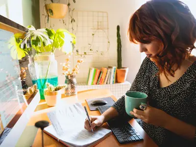 A woman with auburn hair writes in a notebook while holding a green coffee mug at a sunny desk with plants.
