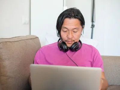 A man in a pink shirt with headphones around his neck studies at a laptop on a sofa.