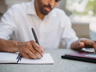 A bearded man in a white shirt writes in a notebook next to a tablet.