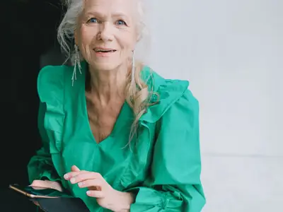 A smiling older woman with silver hair, in a green blouse, uses her smartphone.