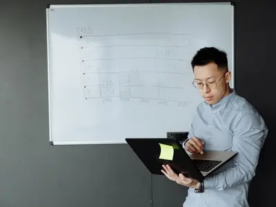 A man holding a laptop stands beside a whiteboard.