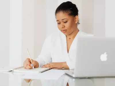 A woman in a white blazer writes notes at a desk beside her laptop.