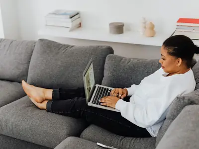 A woman relaxes on a gray sofa, typing on a laptop on her lap.